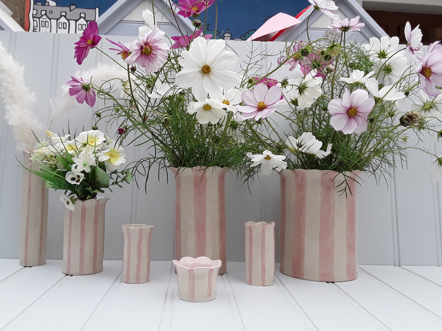 Sea Bramble Ceramics - Handmade, Large, Stoneware vase from their Daisy vase range. With pink stripes and Sea Brambles' signature scalloped top. This vase is ideal for fuller flower arrangements. Shown from left to right are - A tall Sea Lavender vase, a Daisy vase, a Sweet Pea vase, a Tealight holder (front), a Large Daisy vase, a Short Sea Lavender vase and an Extra-Large Daisy vase.
