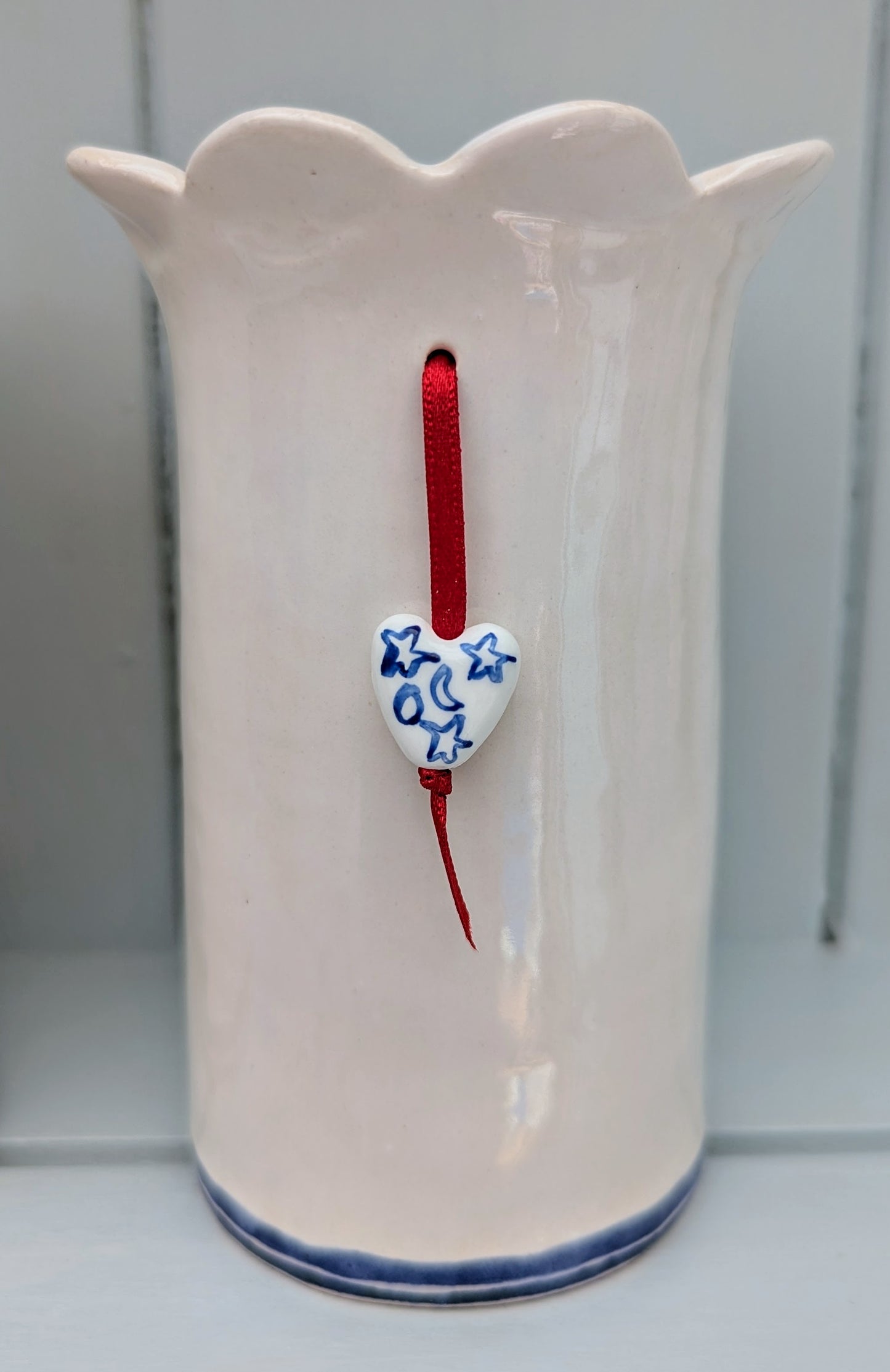 Nautical white ceramic vase with blue edge and a decorative ceramic charm tied on a red ribbon presented on a pale blue background. Designed and handmade exclusively by Sea Bramble Ceramics.