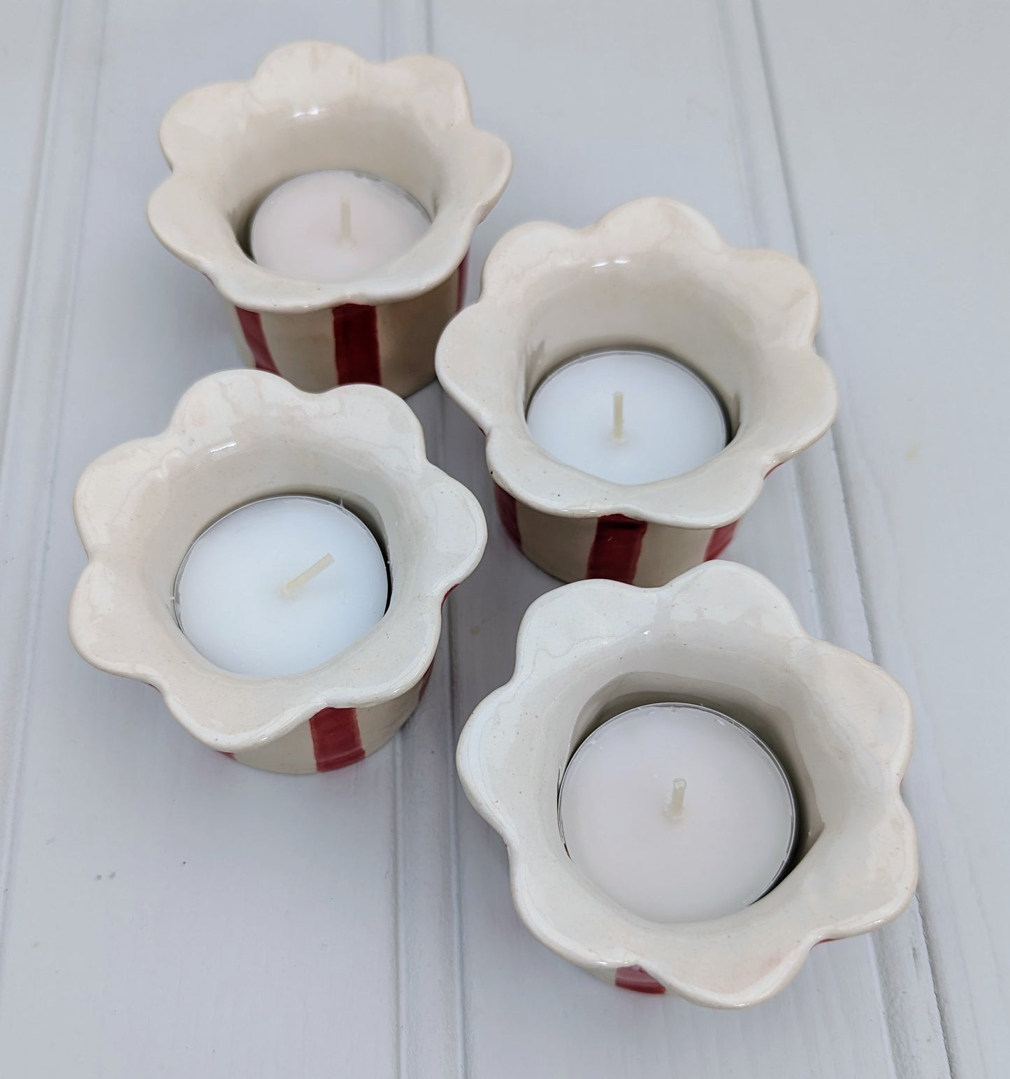 Four ceramic tea light candle holders with a scalloped flower-shaped rim, featuring a cream glaze and red stripes, shown on a white paneled surface.