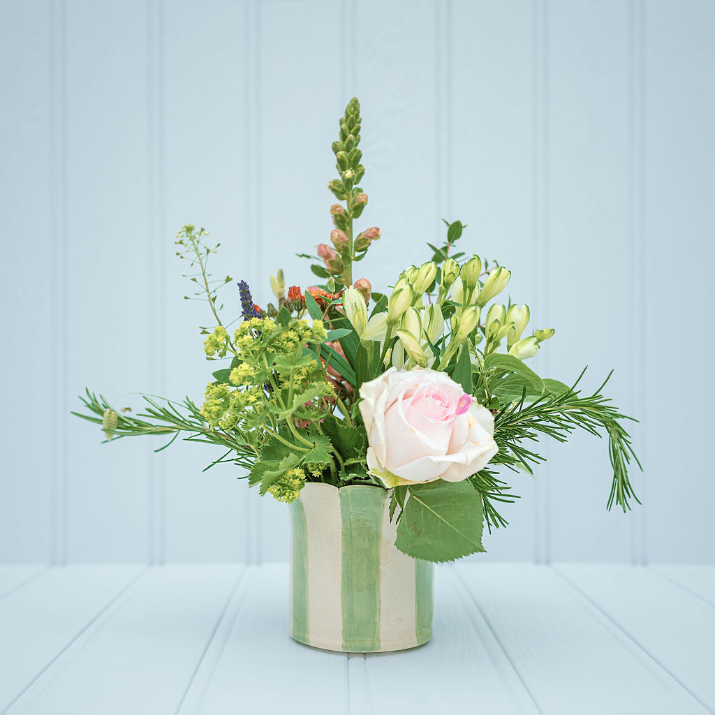 A floral arrangement featuring a pale pink rose, green lily buds, and various green foliage stems in a striped sage ceramic planter, set against a light blue paneled background, all by Sea Bramble ceramics.
