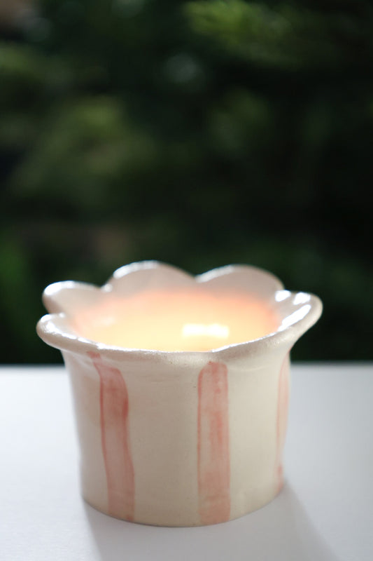 A handmade ceramic tealight holder with a scalloped rim and vertical pink stripes, holding a glowing candle, set against a dark green blurred background. By Sea Bamble Ceramics.