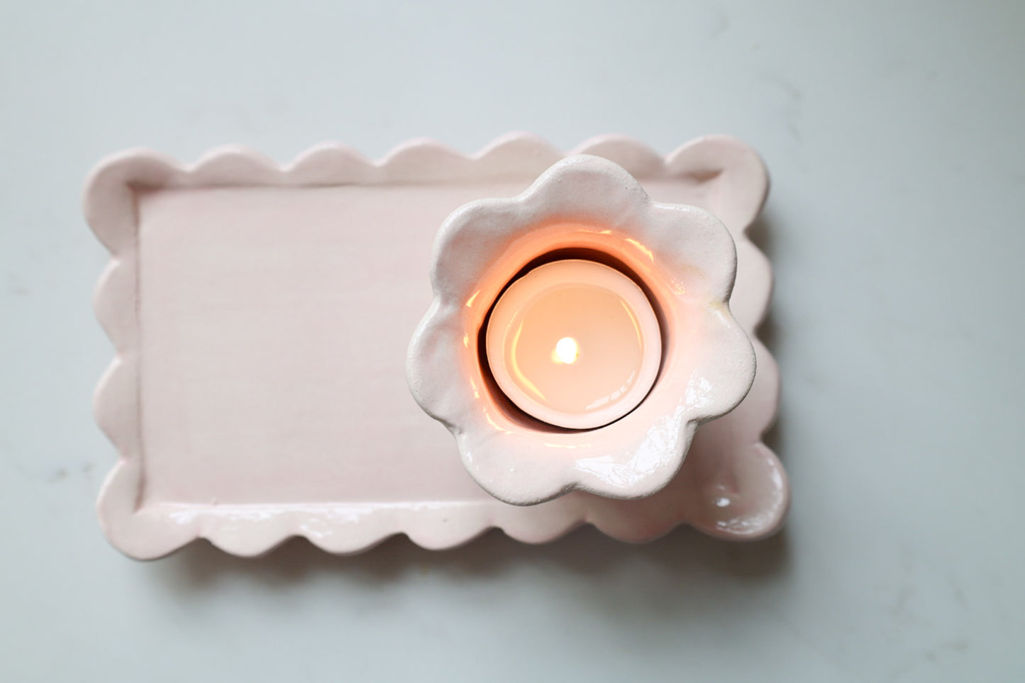 A pink ceramic candle holder with a scalloped rectangular tray and a matching flower-shaped cup holding a lit tealight candle, photographed from above on a light marble surface.
