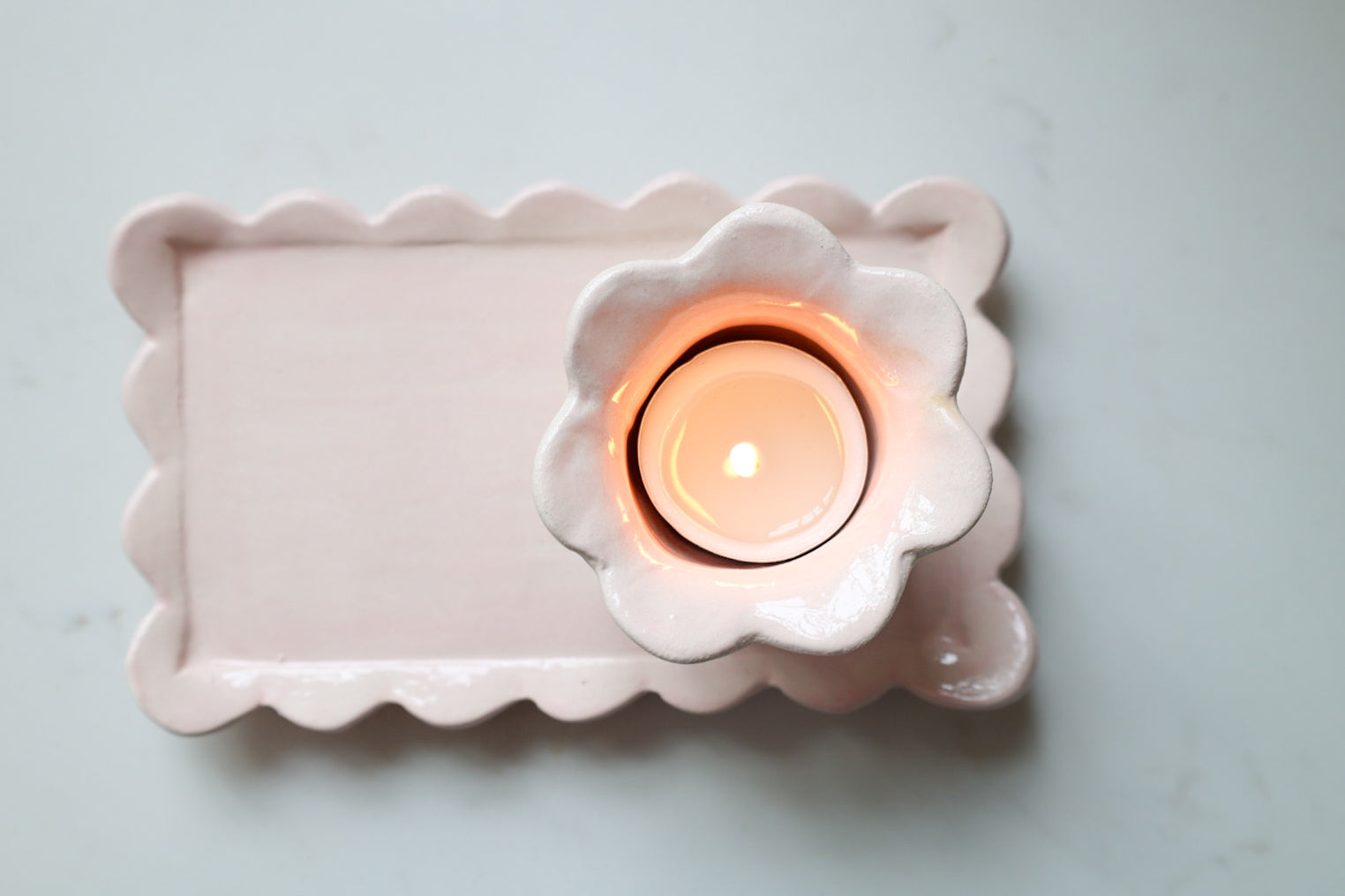 A pink ceramic candle holder with a scalloped rectangular tray and a matching flower-shaped cup holding a lit tealight candle, photographed from above on a light marble surface.
