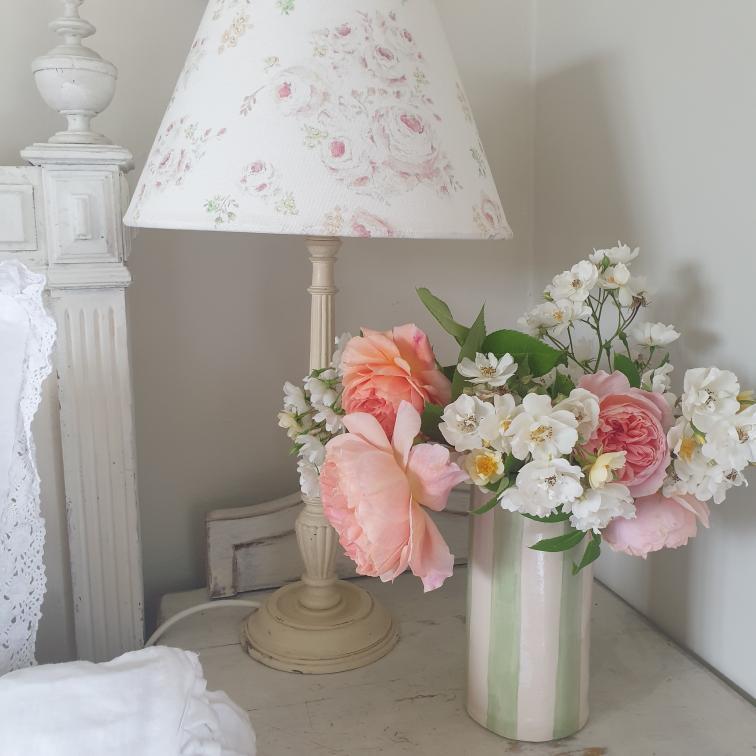 A vintage-style table lamp with a white floral lampshade and cream base sits beside a, Sea Bramble Ceramics, vase of pink and white flowers on a light-coloured bedside table.