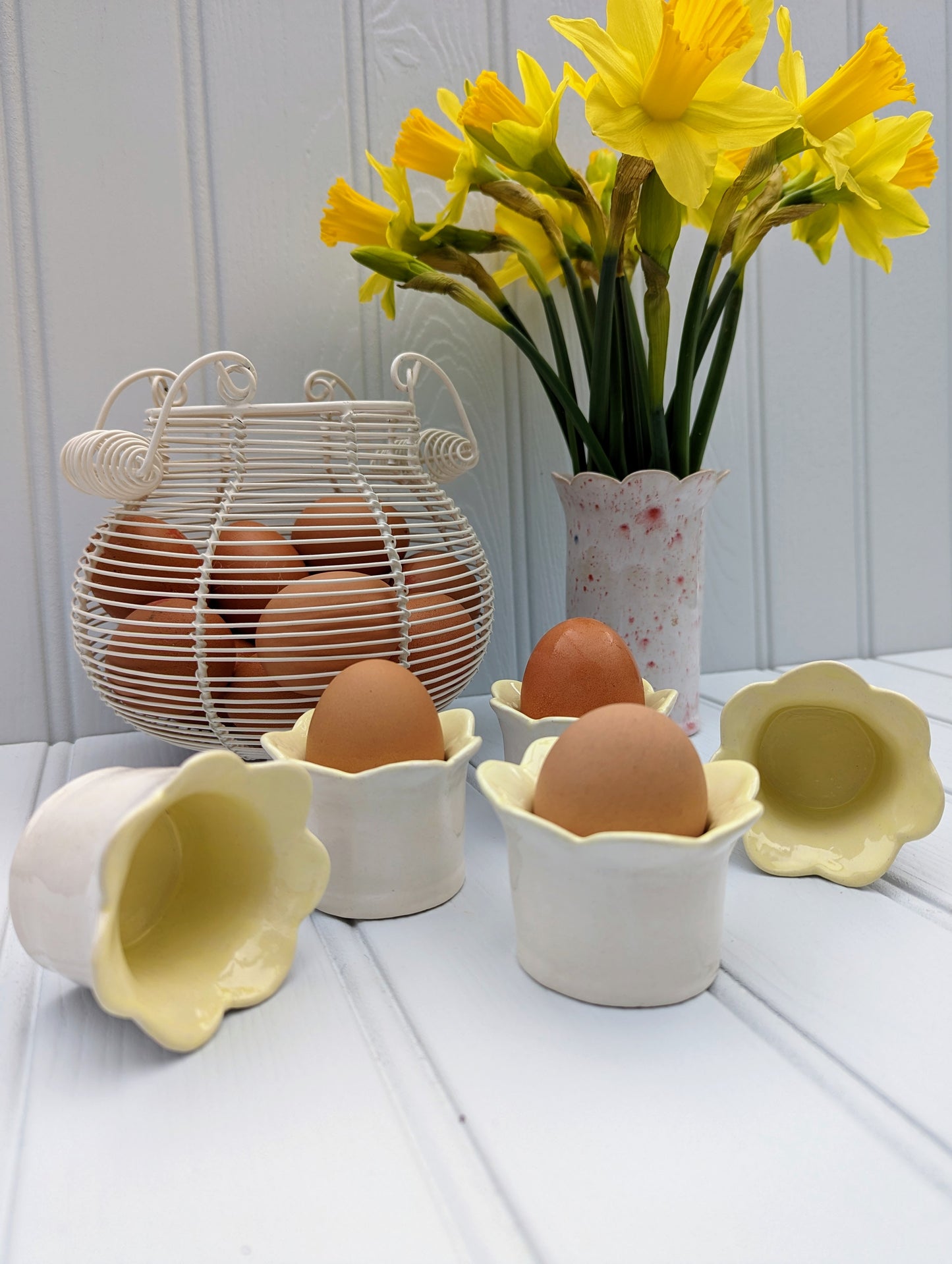 Sea Bramble Ceramics - Handmade Earthenware Daisy egg cups with a scalloped top and glazed with  a white exterior and  yellow interior. Shown here with eggs, egg basket and a beautiful bunch of daffodils in a red speckled Sea Bramble Daisy vase.