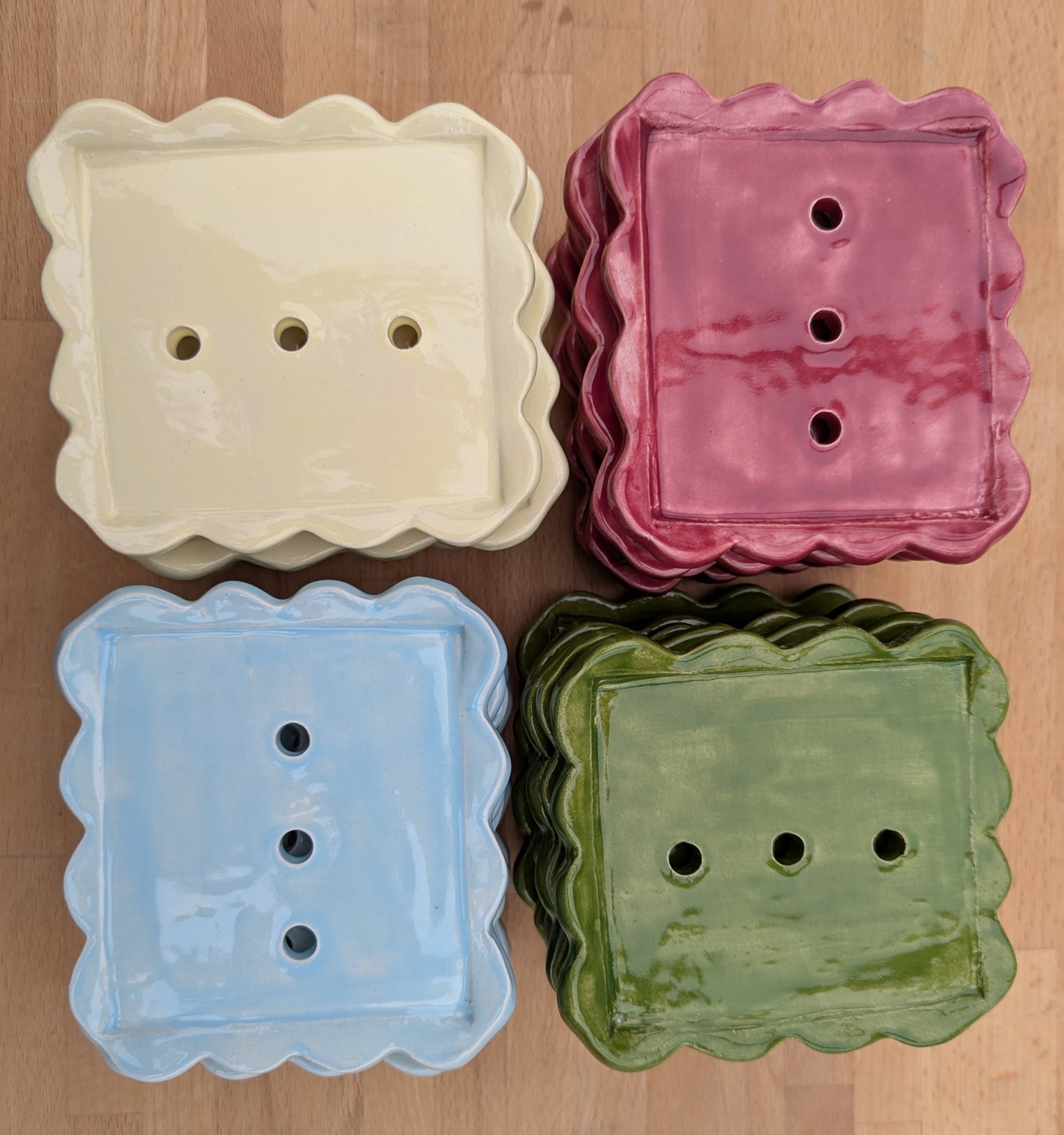 Four square ceramic soap dishes with scalloped edges and three drainage holes, shown in yellow, dark pink, light blue, and olive green, stacked on a light wood background.