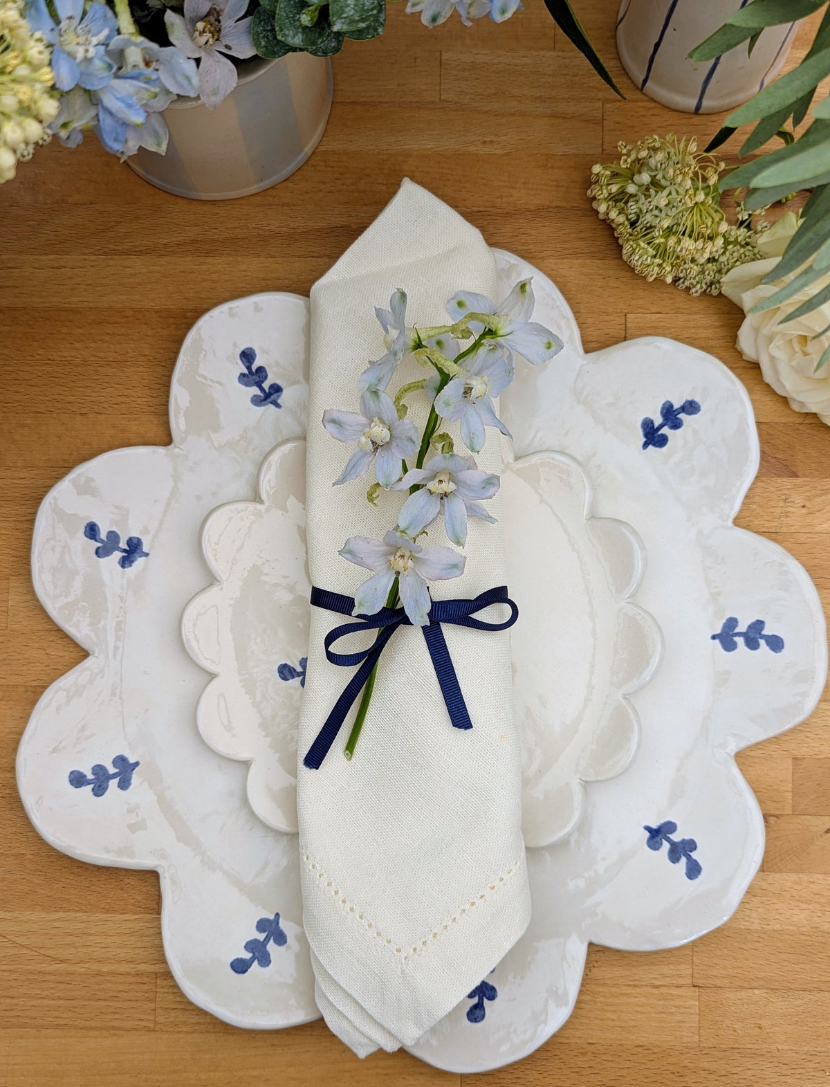 A folded white napkin with a navy blue ribbon bow and a small sprig of pale blue flowers, placed on top of matching white scalloped plates with blue motifs on a wooden table.