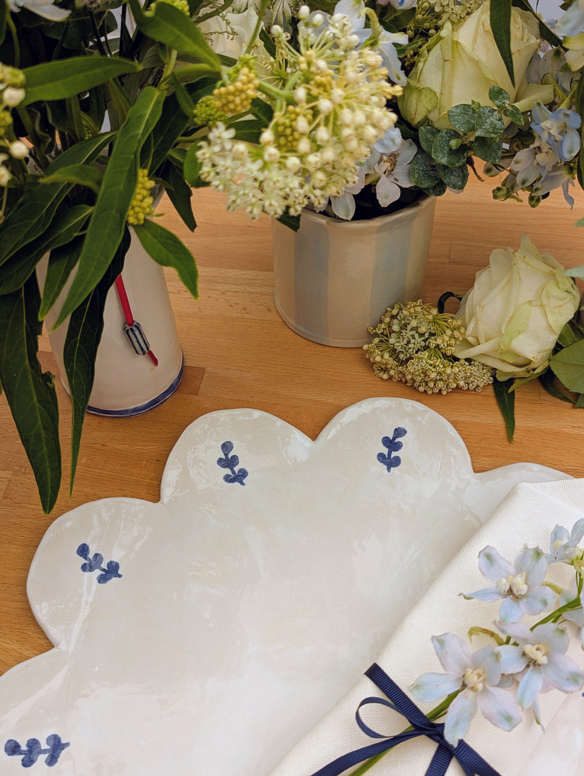 A white cloud-shaped ceramic plate with blue floral motifs, styled on a wooden table alongside white flowers and greenery.