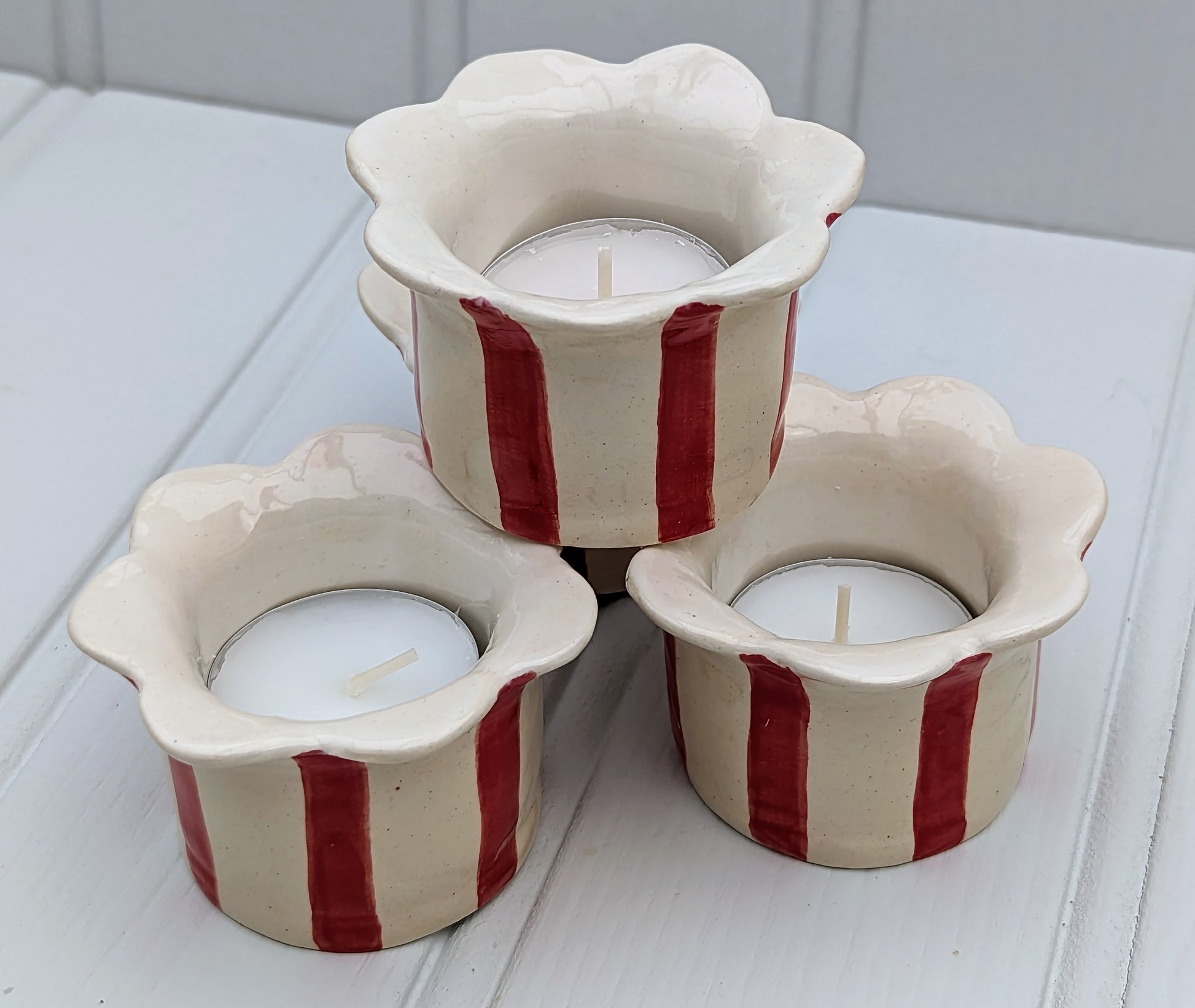 Three ceramic tea light holders with a scalloped flower-shaped rim, decorated with vertical red stripes on an off-white base, arranged on a white wooden surface.
