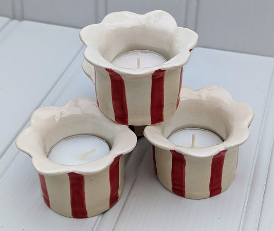 Three ceramic tea light holders with a scalloped flower-shaped rim, decorated with vertical red stripes on an off-white base, arranged on a white wooden surface.